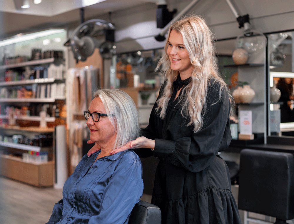 Frisör hjälper kund med naturligt grått hår i salong – professionell hårvård och styling.